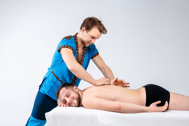 Тематичний масаж і догляд за тілом. Красивий білий чоловік у блакитній формі і борода робить розтягування, лікує, діагностує хлопця з м'язами спини з хорошою фігурою на масажному столі перед білим тлом
 - Фото, зображення