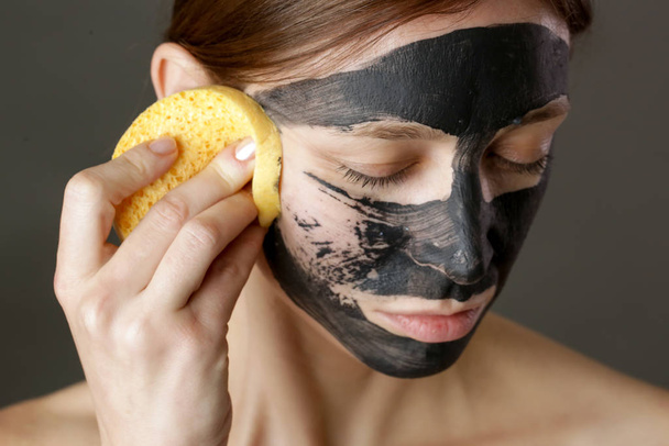 Μάσκα προσώπου από κάρβουνο ή μαύρη λάσπη από πηλό. Όμορφη γυναίκα βγάζει μαύρη μάσκα από το πρόσωπό της, κλείνει το στούντιο. Φυσική αισθητική έννοια. - Φωτογραφία, εικόνα