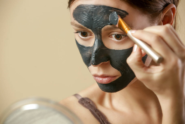 Houtskool masker of zwarte klei modder. Mooie vrouw is het aanbrengen van zuiverende zwarte masker op haar gezicht, close-up studio schot. Home spa natuurlijke cosmetische concept. - Foto, afbeelding
