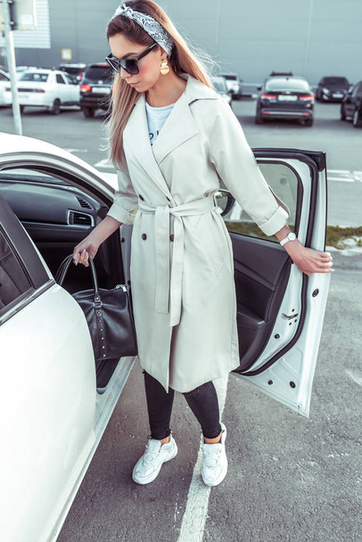 beautiful girl in raincoat, in summer autumn city, goes out and gets into car interior holding bag with purchases from store. Car rental, car sharing. Car parking at mall. - Foto, afbeelding