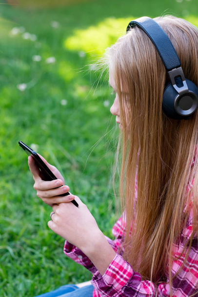 Mooi blond tienermeisje dragen jeans zittend op het gras met koptelefoon en mobiele telefoon - Foto, afbeelding