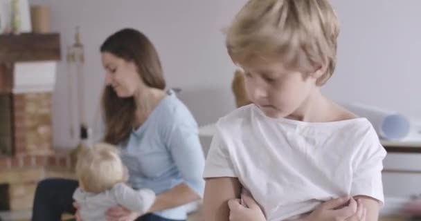 Portrait of upset Caucasian boy with grey eyes looking back at his sister and mother playing, turning back and looking away. Guy in white T-shirt posing at home. Cinema 4k ProRes HQ. - Filmmaterial, Video