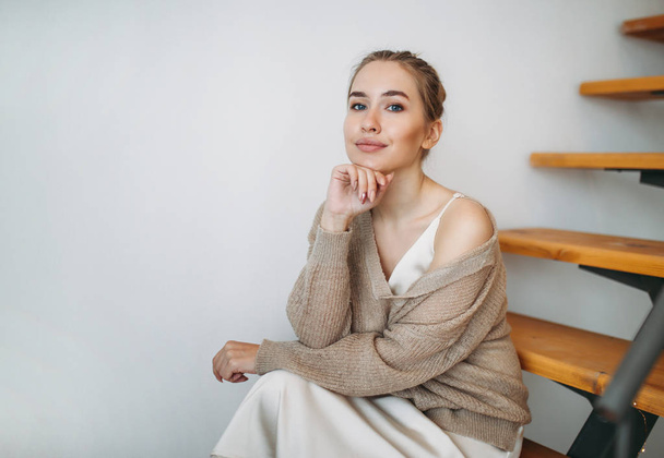 Joven hermosa chica con cabello rubio en vestido de seda beige noche y acogedor cárdigan sentado en las escaleras en casa
 - Foto, Imagen