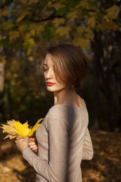 Menina feliz com cabelo curto loiro e um belo sorriso em um vestido bege mantém um buquê de folhas de outono em um belo parque de outono vermelho-ouro
 - Foto, Imagem