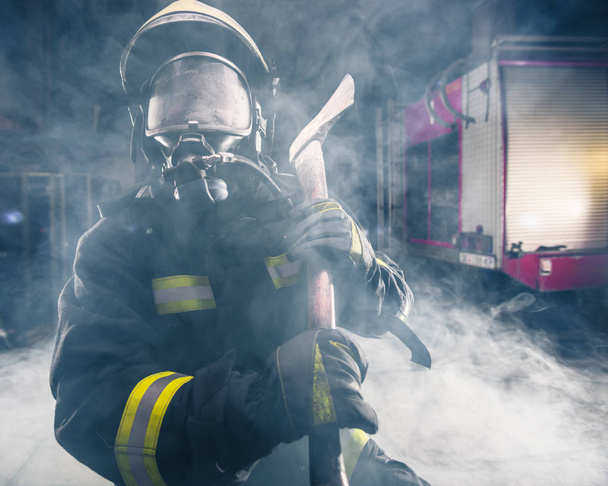 Retrato de una mujer bombero mientras sostiene un hacha y lleva una máscara de oxígeno en el interior rodeado de humo. - Foto, imagen