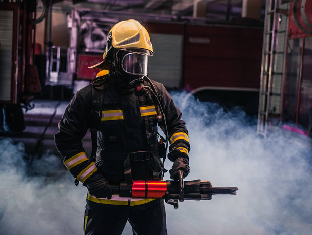 Портрет молодого пожарного, стоящего и держащего бензопилу посреди дыма бензопилы - Фото, изображение