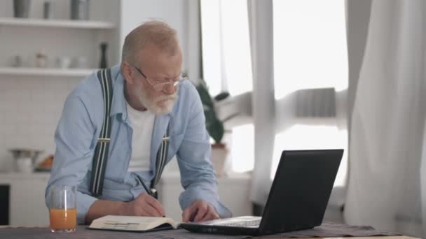 handsome bearded elderly man in glasses working freelance on laptop computer at home leads active modern lifestyle of older people - Felvétel, videó
