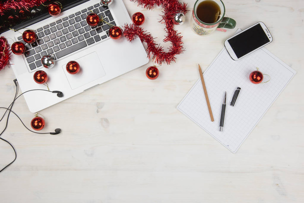 Praca w święta Bożego Narodzenia: płaskie układanie jasnego drewnianego stołu z otwartym aluminiowym laptopem, czerwoną dekoracją, czerwonymi bombkami, świąteczną filiżanką herbaty, smartfonem, notatkami, ołówkiem i długopisem na karteczce - Zdjęcie, obraz