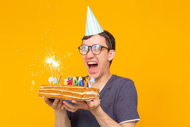 Crazy cheerful young asian guy with glasses holding a burning candle in his hands and a congratulatory homemade cake on a yellow background. Birthday and anniversary celebration concept. - Photo, Image