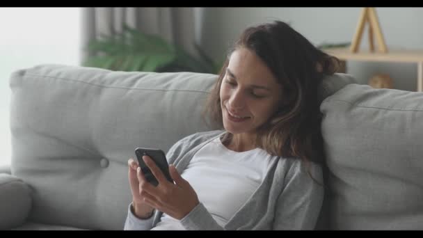Happy young woman relaxing on cozy couch, using smartphone. - Filmati, video