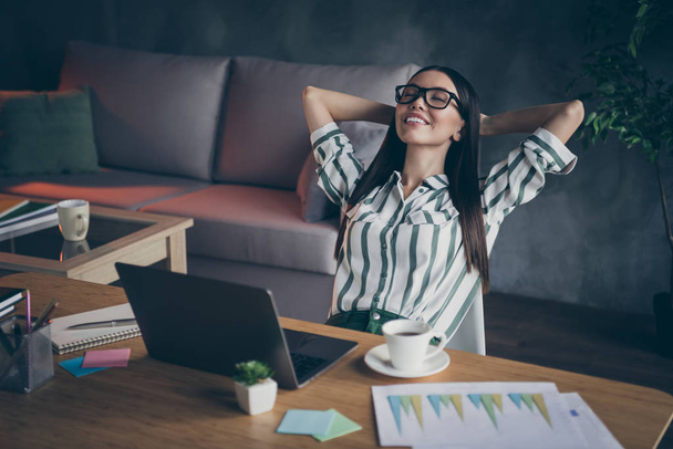 Photo of cheerful relaxing girl having rest at her work place smiling toothily lying on hands behind her head in spectacles - Фото, зображення