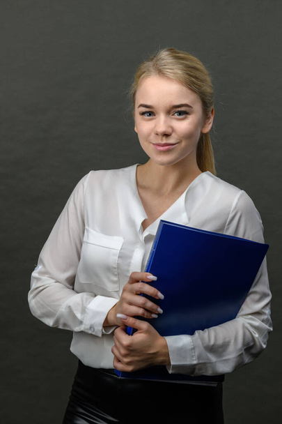 Portrait of young beautiful blonde businesswoman holding blue folder on grey background - Foto, Imagen