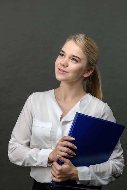 Porträt einer jungen schönen blonden Geschäftsfrau mit blauem Ordner auf grauem Hintergrund - Foto, Bild