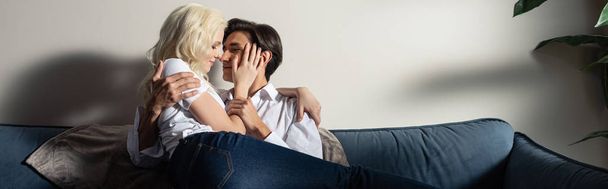 Panoramiczne ujęcie uśmiechniętej młodej pary przytulającej się na kanapie w salonie - Zdjęcie, obraz