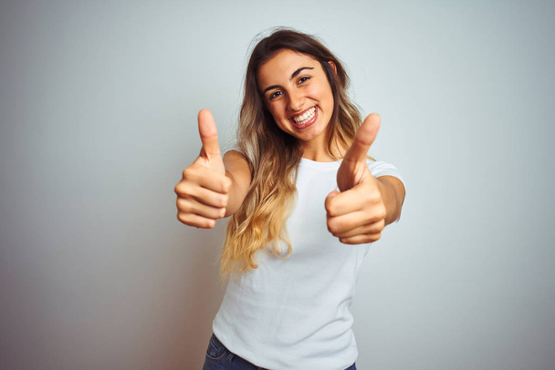Mladá krásná žena na sobě ležérní bílé tričko přes izolované pozadí znamení úspěchu dělá pozitivní gesto s rukou, palce nahoru s úsměvem a šťastný. Veselý výraz a gesto vítěze. - Fotografie, Obrázek