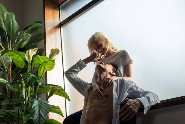 Smiling woman hugging shirtless man beside window in living room - Valokuva, kuva