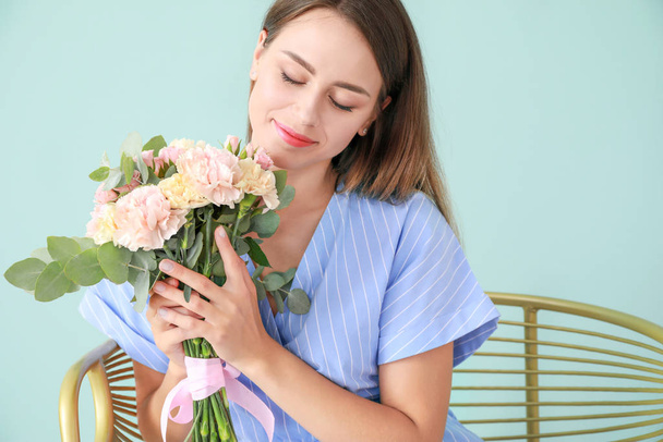色の背景にアームチェアに座っているカーネーションの花の花束を持つ美しい若い女性 - 写真・画像