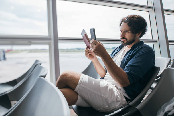Ένας άντρας κάθεται κοντά στην πύλη του αεροδρομίου με ένα τηλέφωνο.. - Φωτογραφία, εικόνα