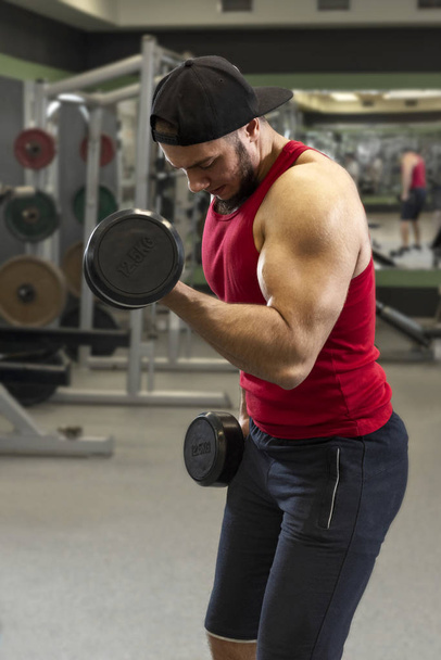 Un joven haciendo ejercicio con pesas. Hombre fuerte entrena bíceps en el gimnasio
. - Foto, Imagen