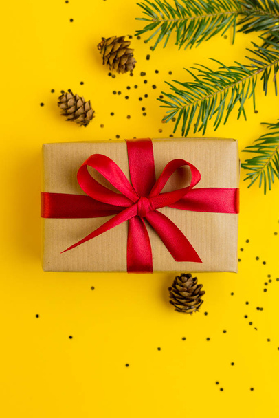 赤いリボンの茶色のギフトボックス。多色のコンフェッティで黄色の背景。フラットレイアウト、トップビュー、コピースペース。モミのクリスマスツリーの枝やコーンとクリスマスのための贈り物 - 写真・画像