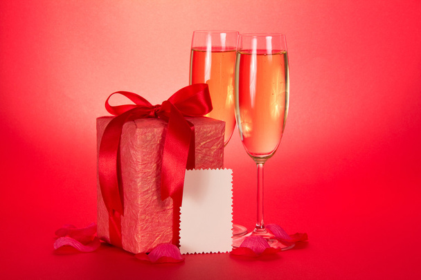 Two wine glasses with champagne, a gift box with a bow, an empty card and petals of roses on a red background - Valokuva, kuva