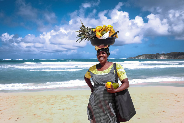 2018.02.24 excursão à praia do Rincon. República Dominicana. Jovem feliz negra afro-americana segurando cesta de frutas na cabeça vendendo frutas frescas à beira-mar com uma praia e ondas de fundo
 - Foto, Imagem