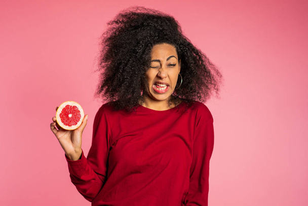 African american girl act ape her face from grapefruit cid. Woman grimaces. Lady with curly hairstyle on pink background. Diet. healthy lifestyle concept. - Photo, image