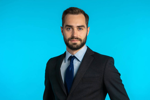 Close up portrait of young successful confident businessman with beard isolated on blue studio background. Man in business suit looking to camera and smiling. Portraiture of handsome guy. - Foto, Bild