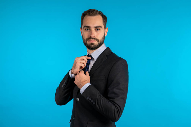 Handsome young businessman corrects his necktie and looking to camera like in mirror. Man on blue studio background. Corporate person in elegant suit correcting his tie. - Φωτογραφία, εικόνα