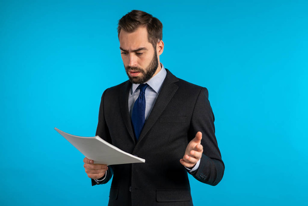 Serious businessman boss isolated on blue background studio. Man is unsatisfied with work of staff. Young mature male office employee in suit jacket with documents, reports or contract - Foto, Bild