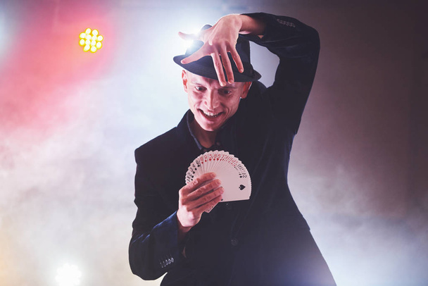 Magician showing trick with playing cards. Magic or dexterity, circus, gambling. Prestidigitator in dark room with fog. - Фото, зображення