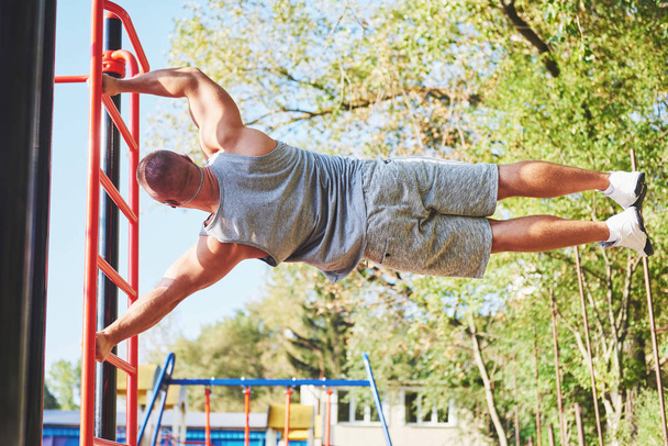 Muscular man with beautiful torso exercising on horizontal bars on a blurred park background. Young man doing pull-ups outdoors. - Фото, изображение