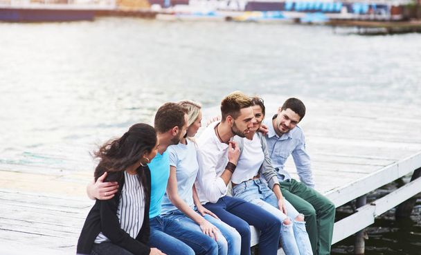 Portrait of joyful group of young people sitting on the edge of the pier, outdoors in nature. Friends enjoying a day on the lake - Foto, Bild