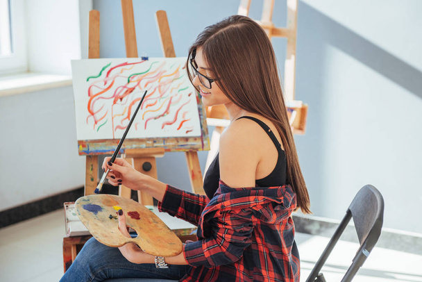 Creative pensive painter girl paints a colorful picture on canvas with oil colors in workshop. - Foto, imagen