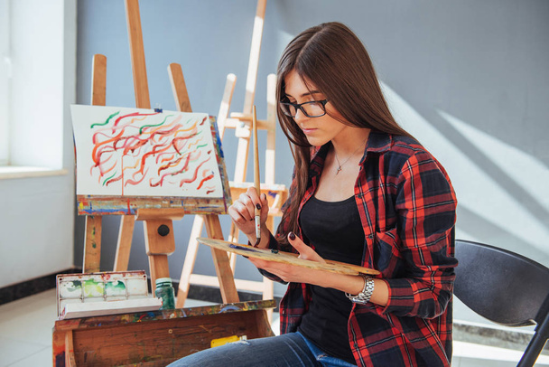 Creative pensive painter girl paints a colorful picture on canvas with oil colors in workshop. - 写真・画像