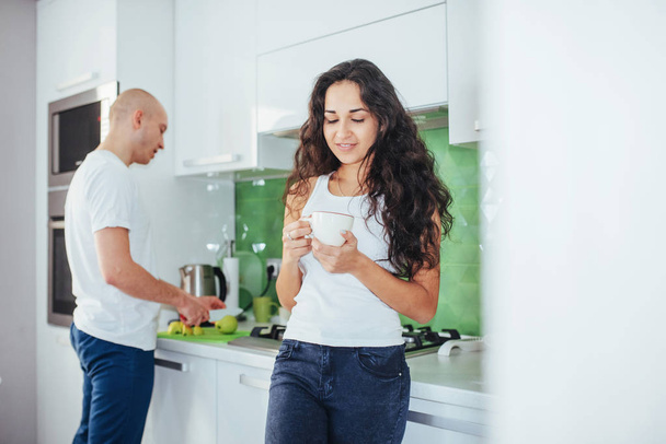 Beautiful young couple is talking, looking at camera and smiling while cooking in kitchen at home. - Photo, image