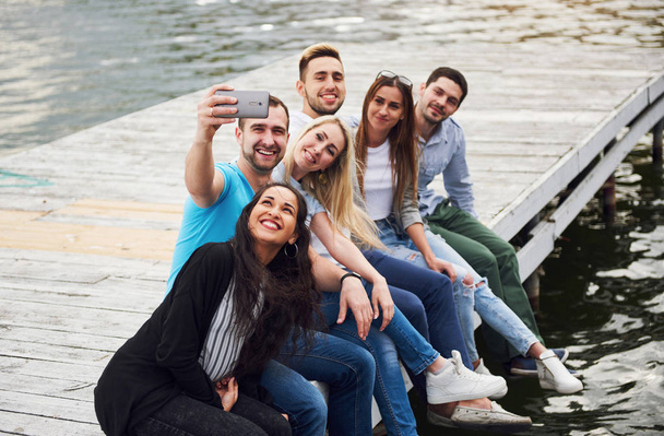 A group of young people sitting on the edge of the pier, and make selfie. Friends enjoying a game on the lake. - Fotografie, Obrázek