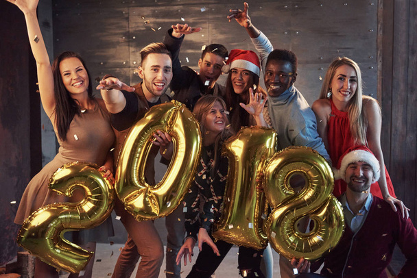 Das neue Jahr 2018 kommt! Gruppe fröhlicher junger multiethnischer Menschen in Weihnachtsmannhüten, die goldfarbene Zahlen tragen und Konfetti auf die Party werfen - Foto, Bild