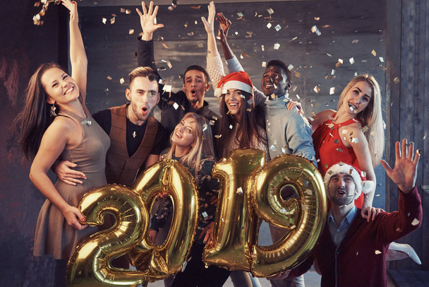 Nowy 2019 Rok nadchodzi! Grupa wesołych młodych wielonarodowych ludzi w kapeluszach Mikołaja niosących złote numery i rzucających konfetti na imprezę. - Zdjęcie, obraz