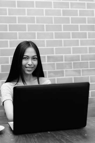 Young happy Asian teenage girl smiling with laptop on wooden table against brick wall - Valokuva, kuva
