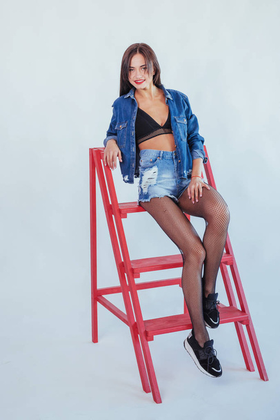 Fashionable brunette beautiful woman posing in studio wearing short jeans, looking at camera. - Фото, изображение