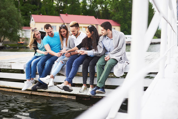 Portrait of happy young friends sitting on a pier at the lake. - 写真・画像