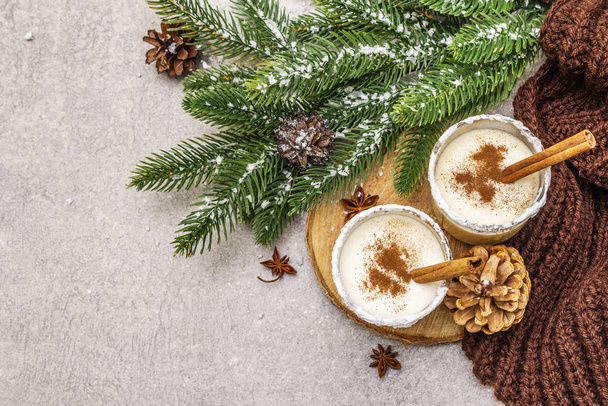 Homemade eggnog with cinnamon in glass. Typical Christmas dessert. Evergreen fir brunch, cones, cozy plaid, artificial snow. Stone concrete background - Zdjęcie, obraz