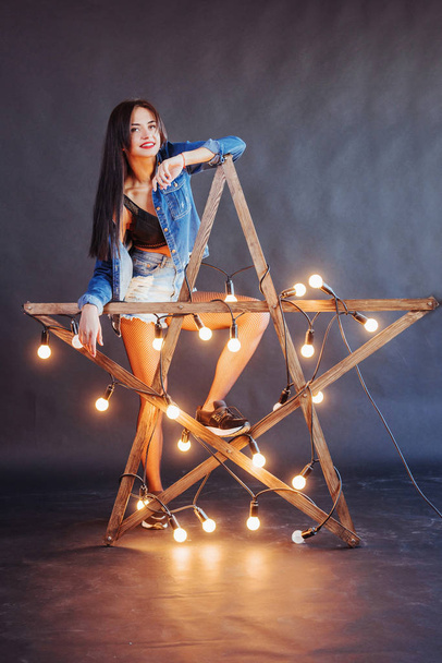 Sexy brunette woman in nice jeans in the studio. Wooden star with lights on a black background. - Foto, imagen