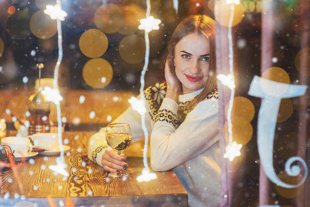 Μια νεαρή όμορφη γυναίκα κάθεται στο καφέ, πίνοντας κρασί. Μαγικό φαινόμενο χιονιού. Χριστούγεννα, νέο έτος, ημέρα του Αγίου Βαλεντίνου, έννοια χειμερινές διακοπές. Η φωτογραφία τραβήχτηκε από το παράθυρο. - Φωτογραφία, εικόνα
