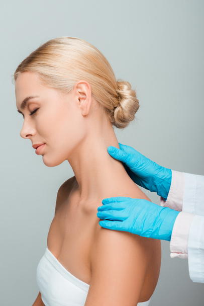 abgeschnittene Ansicht einer Kosmetikerin in blauen Latex-Handschuhen, die den Hals einer Frau mit geschlossenen Augen berührt, isoliert auf grau  - Foto, Bild