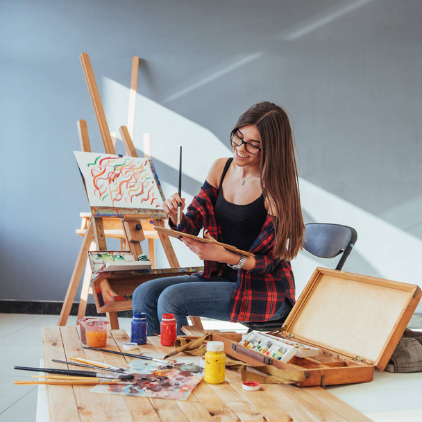 Creative pensive painter girl paints a colorful picture on canvas with oil colors in workshop. - Fotó, kép