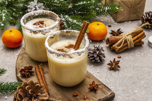 Eggnog with cinnamon and nutmeg for Christmas and winter holidays. Homemade beverage in glasses with spicy rim. Tangerines, candles, gift. Stone concrete background - Foto, afbeelding