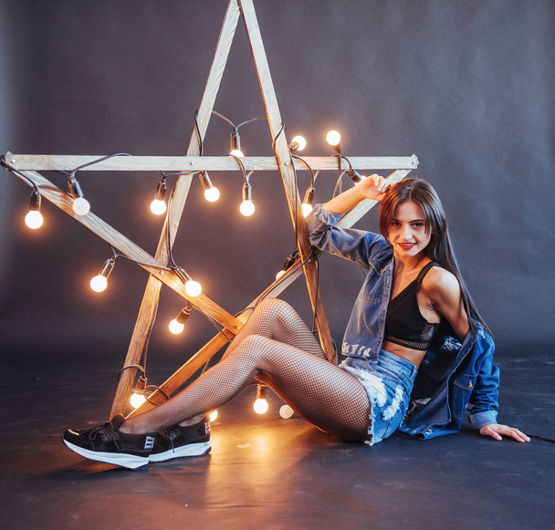 Sexy brunette woman in nice jeans in the studio. Wooden star with lights on a black background. - Фото, зображення