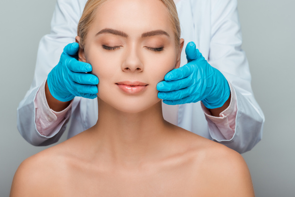 cropped view of beautician in blue latex gloves touching cheeks of naked woman isolated on grey  - Fotoğraf, Görsel